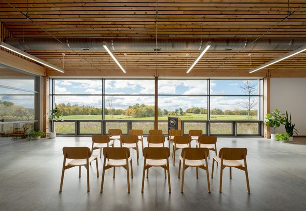 A spacious, bright multi-purpose interior with rows of timber dining or meeting chairs arranged before expansive floor-to-ceiling glazing. The room features a warm exposed timber slatted ceiling, suspended linear LED lights, visible ductwork, polished concrete flooring, and simple white walls. A broad landscaped rural view is visible through the windows, with potted plants adding greenery indoors.