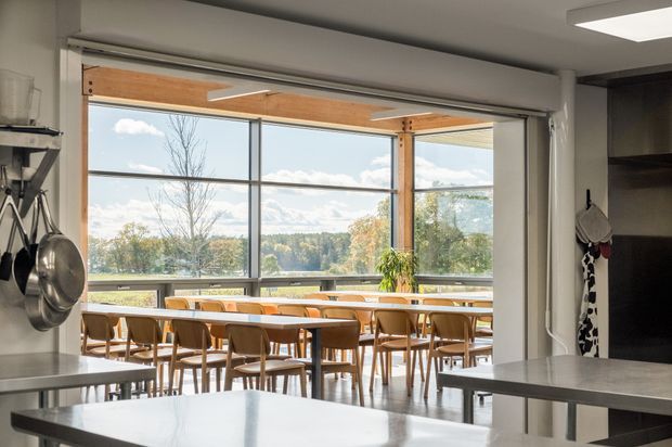 A bright, open-plan dining area viewed from a kitchen, with a long timber dining table and numerous wooden chairs positioned beside expansive floor-to-ceiling glazing. Timber structural beams and posts frame the glazed opening, while a leafy landscape is visible beyond. Stainless-steel kitchen work surfaces and hanging cookware occupy the foreground, with dark built-in cabinetry at right.