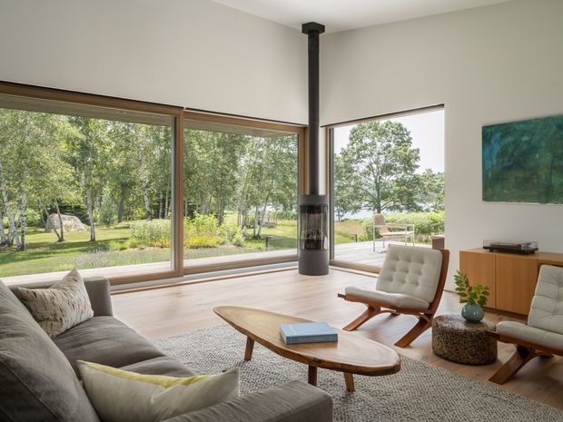 Bright, spacious living room with floor-to-ceiling timber-framed glazing overlooking a landscaped garden. The room features a suspended black cylindrical fireplace, low timber media cabinetry, a large neutral sectional sofa, sculptural wooden lounge chairs, an organic-shaped timber coffee table, and a textured rug. The restrained palette and abundant natural light create a calm, warm mid-century-inspired interior.