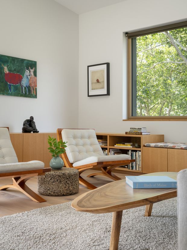 A bright, carefully styled living room with white walls, warm timber built-in cabinetry and shelving, and a large timber-framed window overlooking leafy greenery. Two cream tufted lounge chairs with sculptural wooden frames face a low organic-shaped wood coffee table. A textured cylindrical stone-like side table, small plant, framed artwork, sculpture, books, and a pale rug add understated mid-century character.