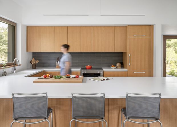 Bright contemporary kitchen viewed from behind a large white island, with warm timber cabinetry, a dark horizontal-tile splashback, integrated appliances, and three metal-framed bar stools. Large windows and a glazed door bring in abundant natural light. The island includes a sink and preparation area, while a person is motion-blurred near the cooktop.