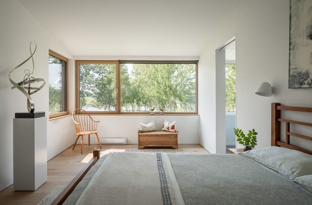 Bright, minimalist bedroom with large timber-framed corner windows overlooking leafy greenery. The room contains a wooden bed with pale bedding, a sculptural artwork on a white plinth, a timber spindle chair, a small wooden chest, a wall-mounted reading light, and a potted plant. The restrained palette and natural light create a calm, airy atmosphere.