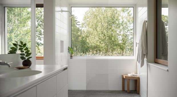 Bright, calm contemporary bathroom with a large freestanding bathtub positioned beneath expansive windows overlooking greenery. White tiled walls, a pale built-in vanity with integrated basin, a frameless shower area, timber-framed mirror, and a small timber stool create a clean, natural interior.