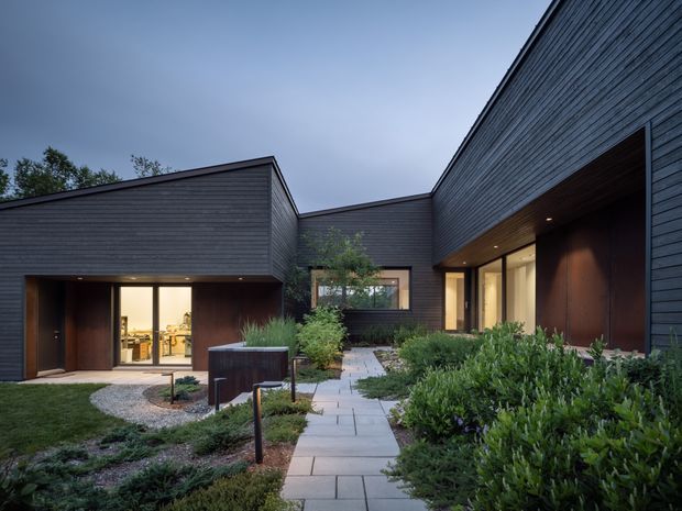 A dusk view of a contemporary courtyard house with dark horizontal timber cladding, low-pitched roof forms, deep sheltered entry recesses, warm interior lighting, and a paved path leading through landscaped planting. Large glazed openings reveal interior spaces, while the courtyard is framed by connected wings.