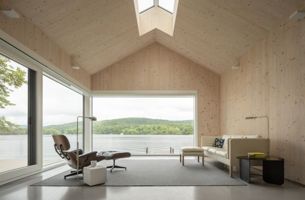 A serene, minimalist living room in a compact gabled pavilion with extensive floor-to-ceiling glazing overlooking a lake and wooded shoreline. The room features pale exposed timber wall and ceiling panels, a central roof skylight, a light neutral sofa and lounge chair, a grey rug, and restrained contemporary furnishings. The gabled ceiling and full-height windows create an airy, nature-connected interior.