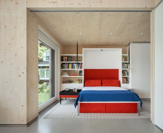 A compact contemporary bedroom viewed through a wide timber-lined opening. The room features a built-in wall bed with vivid red upholstery and a blue blanket, flanked by open bookshelves and white storage cabinetry. Pale timber wall and ceiling linings, large glazing, and a light smooth floor create a bright, streamlined interior.