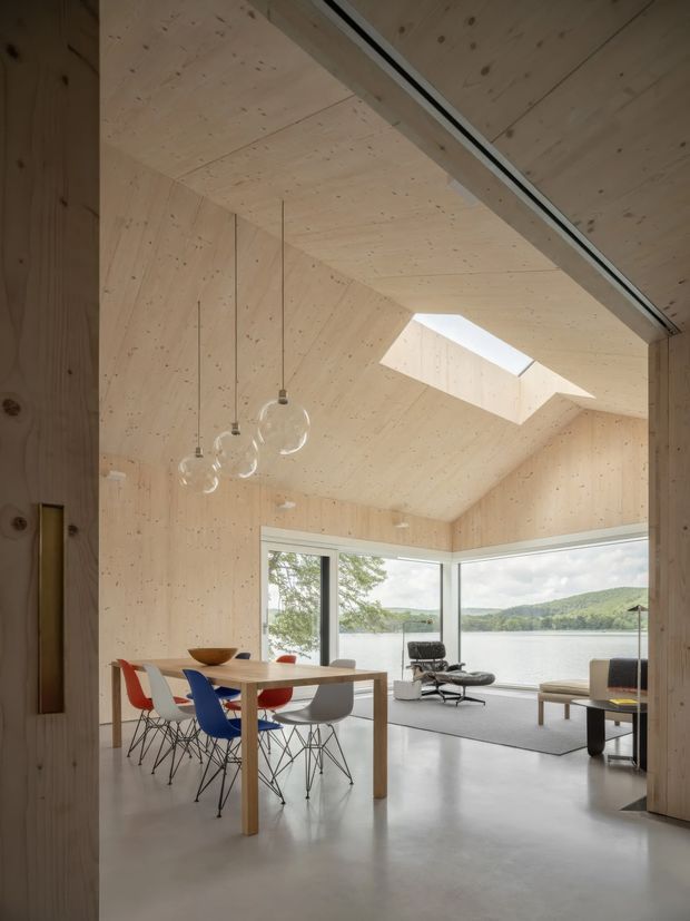 A light-filled open-plan dining and living space clad in pale exposed timber panels. A long wooden dining table with colourful molded chairs sits beneath minimalist pendant lights, while a skylight illuminates the sloped timber ceiling. Floor-to-ceiling corner glazing opens views toward a lake and wooded hills. The room includes a lounge chair, sofa, and low coffee table, with a polished concrete floor and restrained modern furnishings.
