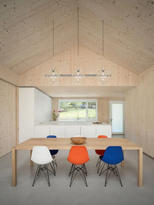 A bright open-plan dining and kitchen interior with a dramatic timber-lined vaulted ceiling and matching timber wall panelling. A long light-wood dining table is surrounded by colourful molded chairs in white, blue, orange and red. Behind it is a minimalist white kitchen with integrated cabinetry, a pale benchtop, a window overlooking greenery, and a frosted-glass door. Three globe pendant lights hang above the table, and the floor is a smooth neutral grey.