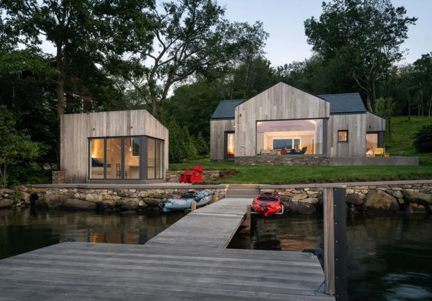 A contemporary timber-clad waterfront retreat viewed from a broad wooden dock. Several compact gabled buildings sit among mature trees on a landscaped, stone-edged shoreline, with warm interior lighting visible through large glazed openings and small watercraft moored at the dock. The composition emphasizes the buildings, dock, and tranquil wooded setting at dusk.