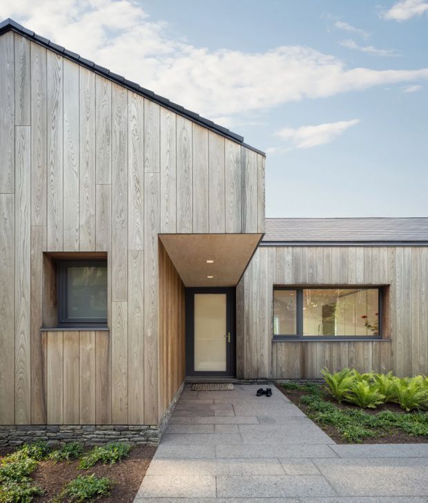 Contemporary single-storey residential facade with vertically arranged weathered timber cladding, dark metal roofing and window frames, and a recessed entry beneath a projecting timber-clad canopy. A paved path leads to the dark-framed front door, with low planting and a stone base wall at the edges. The composition emphasizes clean geometric forms and natural materials.