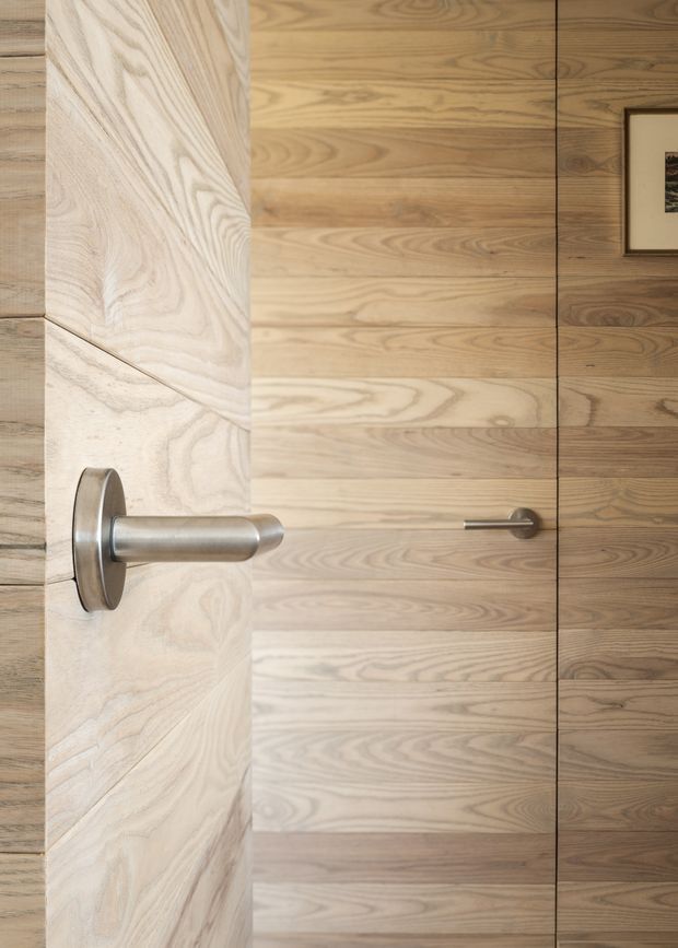 Close-up architectural detail of a flush timber door set within a continuous wood-panelled wall. The door and surrounding panels feature prominent pale natural wood grain, with a minimal brushed-metal lever handle and a small framed artwork partially visible at right.