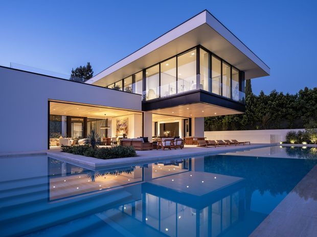 Contemporary two-storey residence photographed at dusk, with strong horizontal flat-roof forms, extensive floor-to-ceiling glazing, warm illuminated interiors, and a large reflective swimming pool in the foreground. The home opens onto a paved pool terrace with outdoor seating and lounge chairs, while landscaped screening surrounds the site.