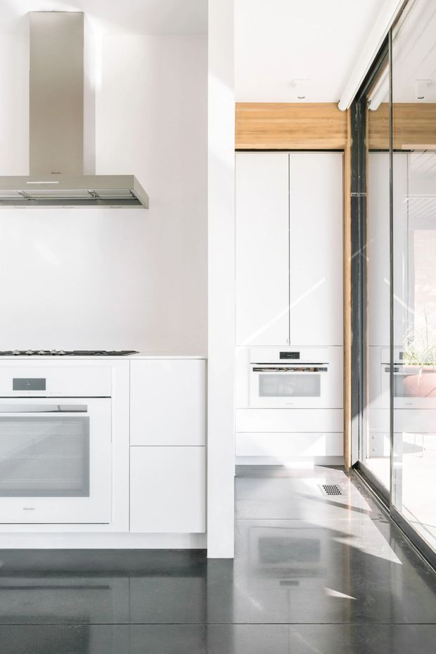 Bright, minimalist modern kitchen with white handleless cabinetry, integrated wall ovens, a built-in cooktop and stainless-steel rangehood. The kitchen opens toward a glazed area with dark-framed windows or doors, warm timber overhead detailing, and highly polished dark grey flooring. Strong natural light creates reflections across the floor and glazing.
