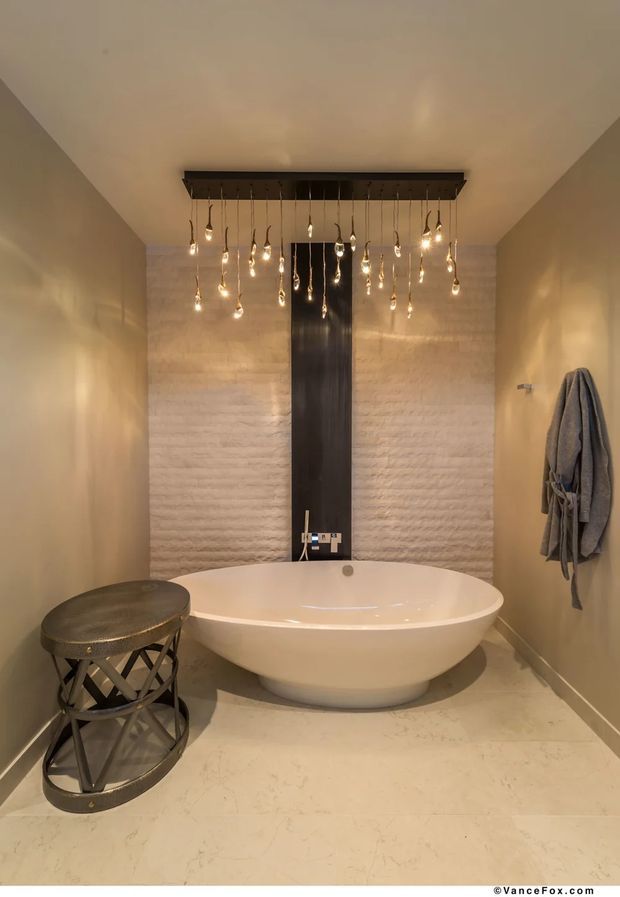 A warm, intimate contemporary bathroom featuring a freestanding oval bathtub centered against a textured light stone wall. A dark vertical feature panel with wall-mounted tapware provides contrast, while numerous exposed pendant bulbs hang from a rectangular ceiling-mounted fixture. Pale stone flooring and walls create a neutral palette, with a dark metallic side stool and grey robe adding functional details.
