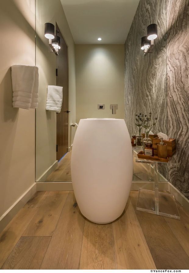 A compact, warmly lit bathroom featuring a freestanding sculptural white bathtub, timber-look plank flooring, a large wall mirror, dark contemporary wall sconces, and a dramatic grey-brown wood-grain patterned feature wall. A clear acrylic side table holds bathroom accessories beside the tub.