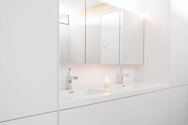 A bright, minimalist bathroom vanity with a long white benchtop, two integrated basins and contemporary chrome tapware. Mirrored wall cabinets span above the vanity, while large white wall and cabinetry panels create a seamless, uncluttered appearance. A small candle provides a warm accent.
