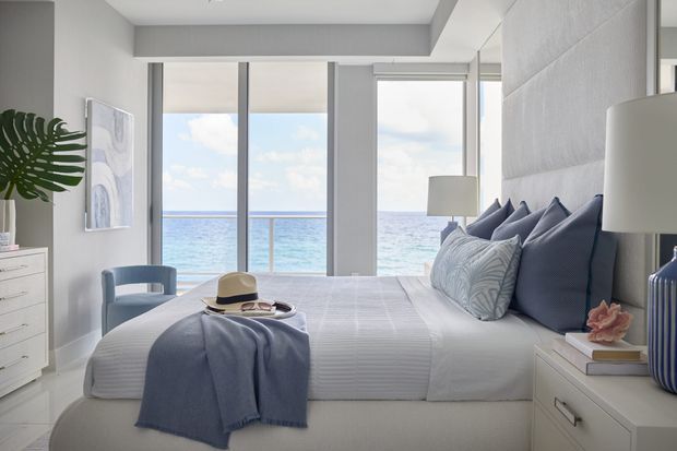 Bright coastal bedroom with a neatly made upholstered bed facing floor-to-ceiling ocean-facing windows. The room features white and light grey finishes, navy and blue bedding accents, white bedside cabinetry, a blue armchair, a large table lamp, and minimal coastal decor.