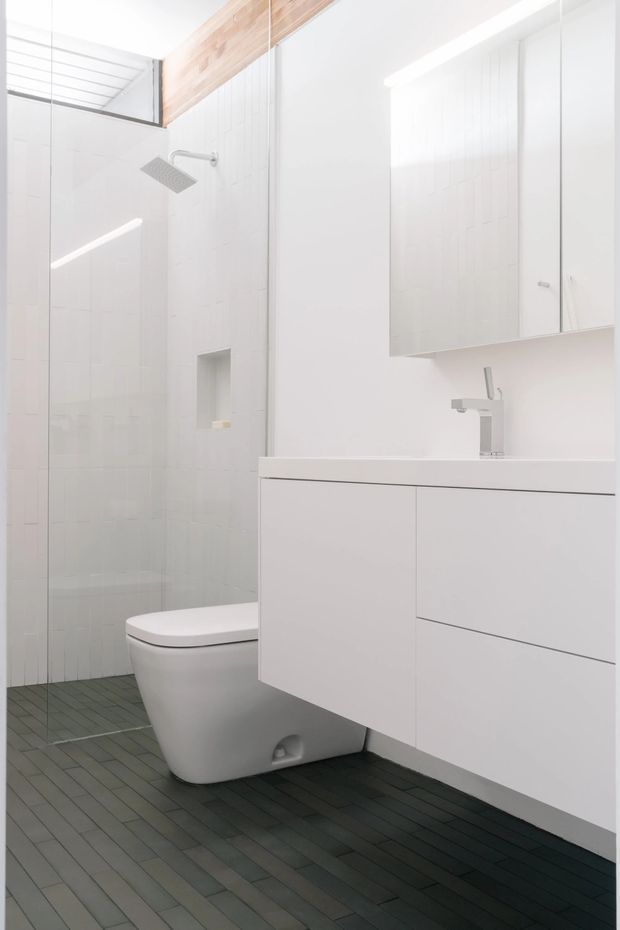 Bright minimalist bathroom with a frameless glass walk-in shower, white tiled walls, wall-mounted white vanity cabinetry, integrated basin and chrome tapware. A compact white toilet sits beside the shower. The floor is finished in narrow dark grey-green rectangular tiles, with a warm timber ceiling or bulkhead accent above the shower.