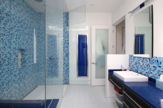 Bright contemporary bathroom featuring a large frameless glass shower enclosure, blue mosaic-tiled walls and splashback, white floor and walls, and a dark floating vanity with a white basin and blue countertop. A frosted-glass door and large mirror complete the clean, high-contrast design.