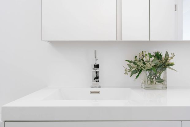A close-up of a minimalist white bathroom vanity featuring a broad integrated rectangular basin, wall-mounted chrome tapware, and simple white overhead cabinetry. A clear glass vase with leafy greenery adds a natural decorative accent. The space is bright, clean, and highly restrained in its detailing.