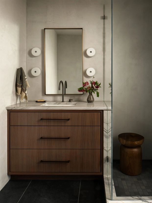 Refined contemporary bathroom vanity with dark timber drawer cabinetry, a pale stone benchtop and integrated basin, framed mirror, sculptural wall lights, and a clear glass shower enclosure. Dark floor tiles and a wooden stool create a warm, moody contrast against the textured light plaster walls.