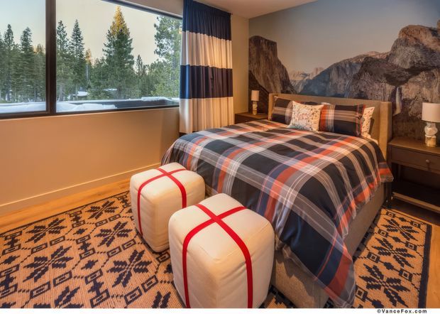 A warmly lit bedroom with a large window overlooking a forested, snowy landscape. The room features a bed dressed in a bold navy, red, grey, and white plaid duvet with coordinating pillows, two white ottomans with red strap details, dark bedside tables, a patterned geometric area rug, striped curtains, timber flooring, and a dramatic mountain mural or photographic wall covering behind the bed.