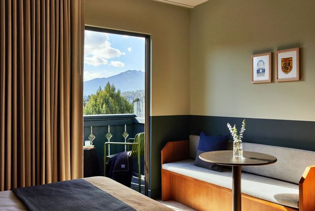 A warm, compact contemporary sitting area adjoining a bedroom, with a built-in L-shaped banquette upholstered in light grey and framed by warm timber cabinetry. A round pedestal table sits in front, while dark lower wall panelling contrasts with olive-beige upper walls. Full-height brown curtains frame a glazed balcony door, providing views toward a mountain landscape. Framed artwork and a small vase add restrained decorative detail.