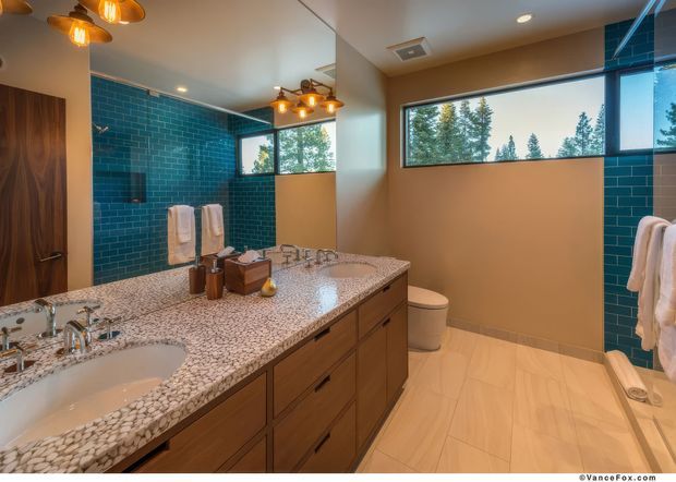 A spacious contemporary bathroom with a long double vanity, warm wood cabinetry, and a pebble-patterned countertop. Teal glazed subway tile forms an accent wall, complemented by light neutral wall and floor finishes, a freestanding toilet, large horizontal windows, and warm pendant lighting. A large mirror reflects the vanity and lighting.