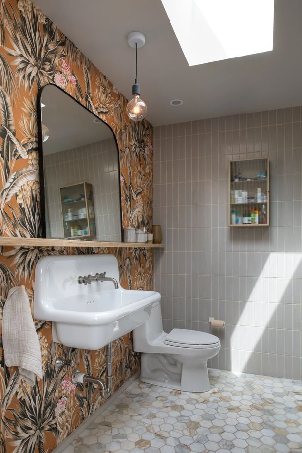 A bright, compact bathroom with a vintage-inspired yet playful design. The room features a wall-mounted white basin beneath a large rounded-rectangle mirror, a white toilet, patterned floral botanical wallpaper on one wall, pale grey rectangular wall tiles, and a hexagonal mosaic tile floor. A pendant globe light hangs beside a large skylight, while recessed shelving provides storage.