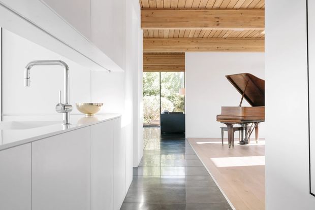 A bright, open-plan interior showing a minimalist white kitchen in the foreground, with a polished concrete walkway leading into a living area containing a grand piano. The space features extensive warm timber ceiling boards and exposed beams, full-height glazing, pale timber flooring, and clean white cabinetry with a sculptural chrome tap and integrated sink.