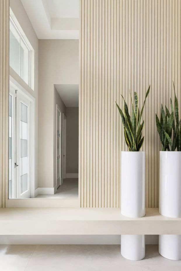 Bright minimalist interior entryway with a tall vertical timber-slat feature wall, white integrated console shelf, matching cylindrical planters, and floor-to-ceiling glazed doors. A hallway recedes into the background, while the neutral palette and abundant natural light create a clean, refined atmosphere.