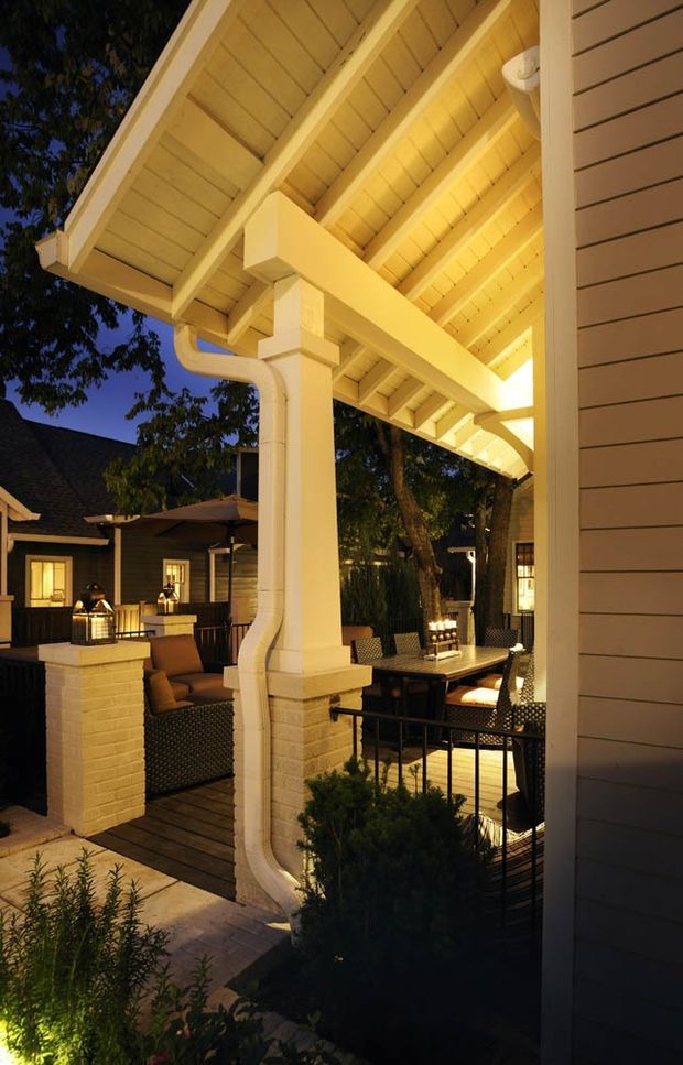 A warmly lit covered outdoor entertaining area at dusk, viewed beside a light-coloured weatherboard-clad house. The structure features a timber-lined pitched roof with exposed rafters, substantial square posts, decorative brackets, white gutters and downpipes, and a timber deck. Outdoor lounge and dining furniture sit around a table, with brick-faced columns, black metal balustrading, planting, and neighbouring house elements visible in the background.