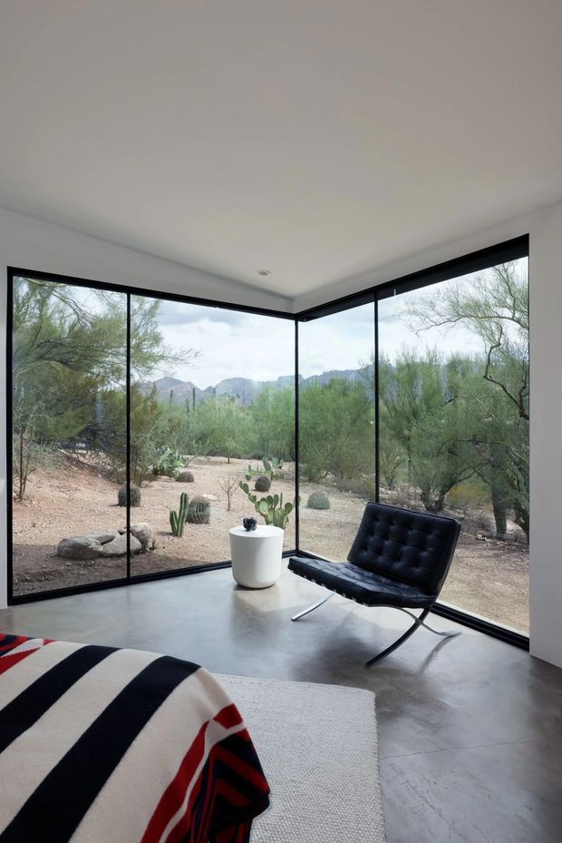 A minimalist living room with floor-to-ceiling corner glazing overlooking a desert landscape. The room has smooth white walls and ceiling, a polished grey concrete floor, black-framed windows, and a distinctive black leather sling chair. A small white planter sits near the glazing, while a striped throw is draped over the foreground seating.