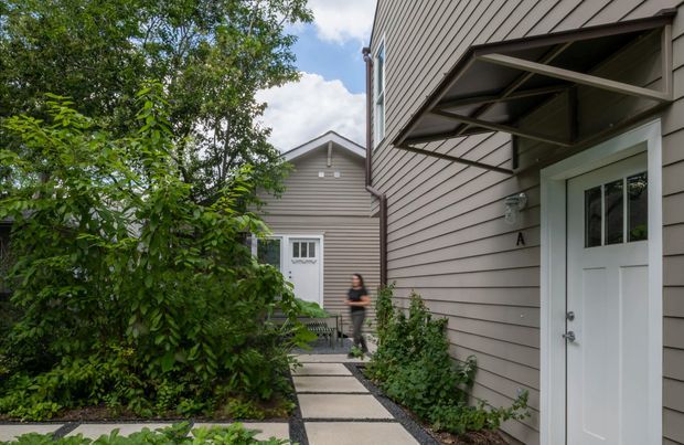 A landscaped pedestrian walkway between two attached or closely spaced light-grey weatherboard buildings. The foreground building has a white entry door beneath a projecting metal awning, while the path leads toward another gabled building with a glazed door. Dense shrubs and small garden beds soften the compact residential-style exterior setting.