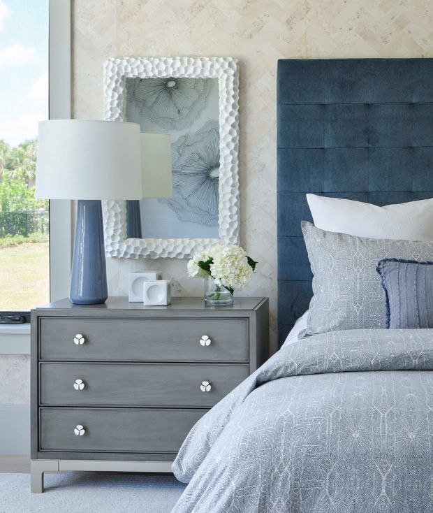 A softly styled bedroom vignette featuring a tall upholstered navy-blue channel-tufted headboard, pale patterned bedding, and a grey three-drawer bedside chest. A blue ceramic table lamp, framed botanical artwork, and a small vase of white flowers sit beside a large window. The room has a calm, refined coastal feel with pale textured wall covering and cool blue-grey tones.