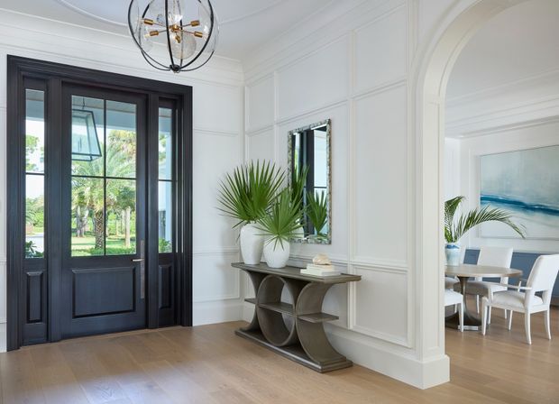 Refined residential entry hall with white decorative wall panelling, a dark timber glazed front door, warm timber flooring, and a sculptural console styled with potted palms and books. A large arched opening leads to a bright dining room with white furniture and coastal artwork. The space has traditional architectural detailing with a fresh, contemporary coastal feel.