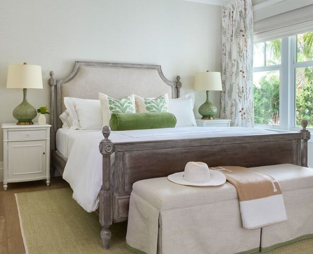 A bright, refined bedroom featuring a substantial upholstered and dark wood four-poster-style bed, white bedding, pale green walls, matching bedside tables with green glass lamps, patterned botanical curtains, and a large upholstered bench at the foot of the bed. A large window provides abundant natural light and views of greenery.