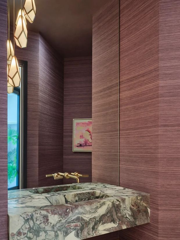 A compact, dramatic powder room featuring dark burgundy-brown textured wall panelling, a large marble vanity with an integrated rectangular basin, and a wall-mounted brass faucet. Geometric pendant lights hang near a narrow window, while a framed floral artwork adds a soft pink accent. The space has a richly layered, luxurious finish.