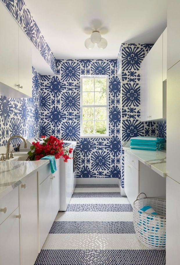 Bright, narrow laundry room with white cabinetry and countertops, a deep utility sink, patterned blue-and-white wall covering, and a striped mosaic tile floor. A central window provides natural light, while a small ceiling fan and laundry baskets reinforce the utility-room function. The room has a playful, bold, highly decorative character.