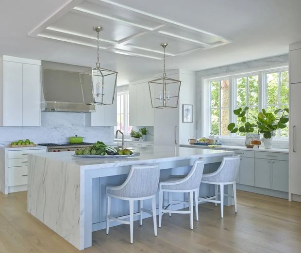 Bright, spacious white kitchen with a large marble-look island, integrated cabinetry, a farmhouse-style sink, and four upholstered bar stools. The room features a decorative coffered ceiling with two lantern pendant lights, pale timber flooring, extensive cabinetry, and large windows overlooking greenery. The styling is refined, airy, and classic with subtle blue-grey accents.