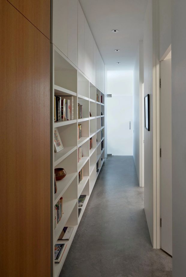A narrow, long interior hallway with full-height white built-in open shelving filled with books along one side, a warm timber wall or door panel at the entrance, white walls and ceiling, recessed downlights, and a smooth grey concrete-like floor. Doorways open off the right side, and bright daylight illuminates the far end. The space has a clean, minimalist built-in storage design.