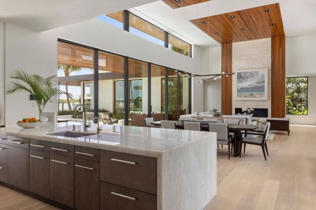 Open-plan contemporary kitchen and dining area with a large white stone island, dark wood cabinetry, floor-to-ceiling glazing, and a dramatic double-height dining space. A warm timber ceiling and vertical timber feature wall frame the dining area, with modern pendant lighting and light wood flooring.
