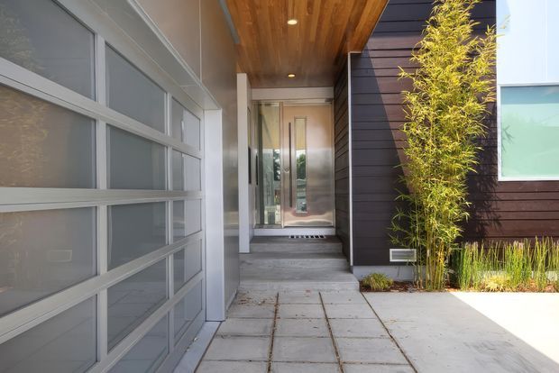 Modern residential entry courtyard viewed from the driveway, featuring a large frosted-glass-panel garage door on the left, concrete slab paving, a recessed front entrance reached by steps, dark horizontal exterior cladding, and a warm timber-lined soffit. Tall bamboo-style planting softens the contemporary facade.