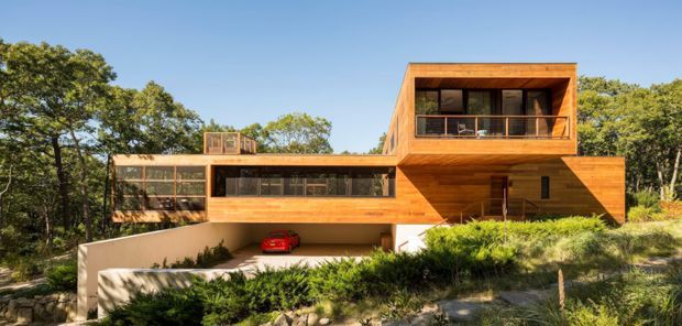 Exterior view of a contemporary multi-level residence set among mature trees and dense landscaping. The home features warm timber cladding, strong cantilevered and geometric volumes, extensive horizontal glazing, a recessed balcony, and an integrated lower-level carport or garage area with a red car visible. The image is photographed from outside and primarily presents the building facade and overall form.