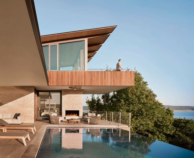 Modern multi-level waterfront residence with broad cantilevered timber balconies, warm brick masonry, expansive glazing, and layered projecting roof planes. The image shows an outdoor entertaining terrace with an infinity-edge pool, lounge seating, built-in fireplace, and views across the water.
