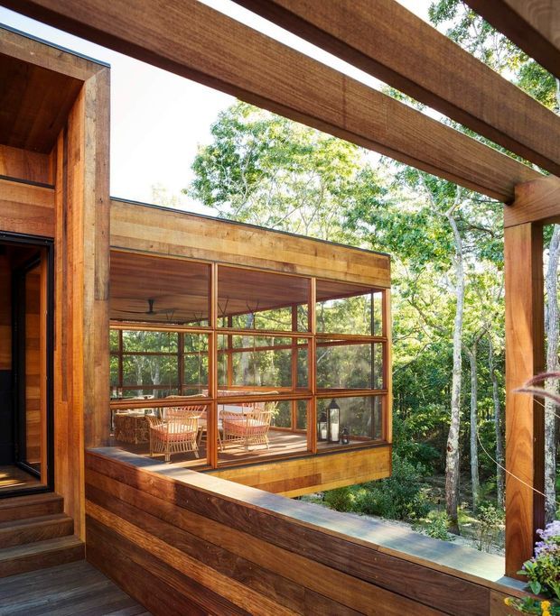A warm timber-clad residential exterior and elevated deck framed by substantial exposed timber beams. A glazed sunroom or living area projects into a leafy wooded setting, with extensive timber-framed glazing, timber flooring, built-in balustrades, and wicker seating visible inside. The image emphasizes the connection between the house and surrounding forest.