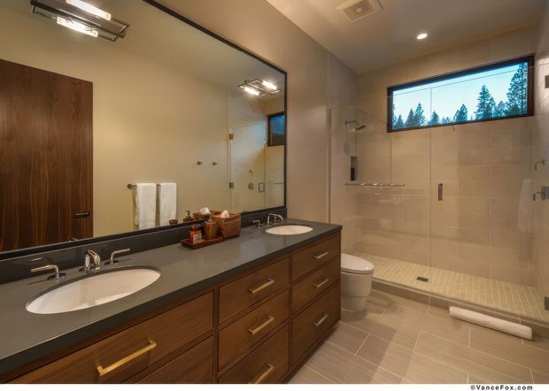 Spacious contemporary bathroom with a long double-sink vanity in dark timber cabinetry, a pale solid-surface benchtop, large wall mirror, and brass-toned hardware. The room includes a walk-in tiled shower with a built-in ledge, handheld and overhead fixtures, a high horizontal window, toilet, recessed ceiling lights, and warm neutral finishes.