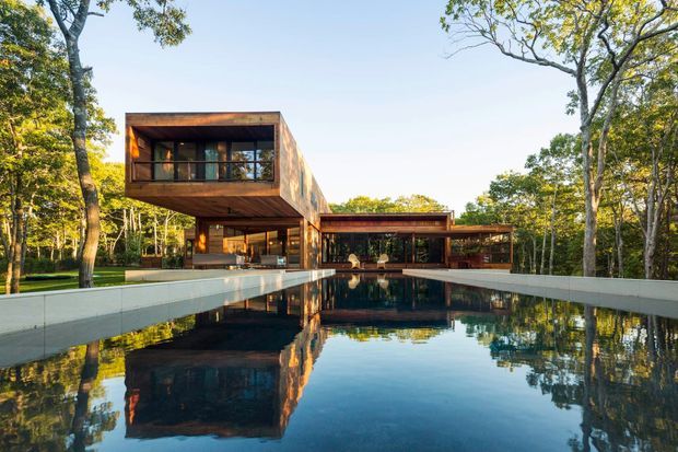A modern multi-level house set within a wooded landscape, photographed across a large reflective swimming pool. The building features cantilevered rectangular volumes, extensive glazing, warm timber cladding, dark structural framing, and broad low-slung roof forms. The pool terrace and surrounding mature trees reinforce the indoor-outdoor connection.