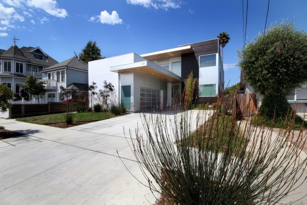 Street-facing view of a contemporary two-storey residence with crisp white rendered walls, dark horizontal timber cladding, broad glazing, a projecting flat-roof canopy and an integrated garage. The house is set in a landscaped suburban lot with a large concrete driveway, low planting and neighbouring traditional homes visible on either side.