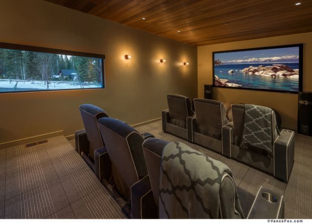 A cozy private home theatre with multiple rows of upholstered reclining cinema seats facing a large wall-mounted screen. The room features warm taupe walls, a richly stained timber-clad ceiling, subtle wall sconces, dark trim, and patterned carpet. A wide horizontal window shows a snowy forest landscape, while the screen displays a bright coastal scene.